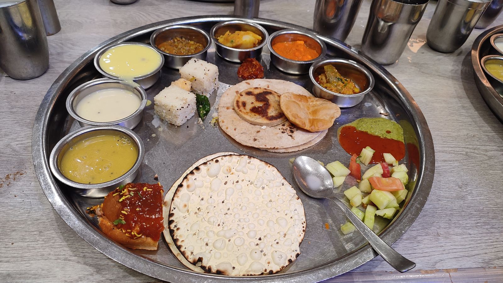 Top-down flat-lay of a traditional steel thali with Gujarati rotli, shaak, dal, and rice on a floral tablecloth